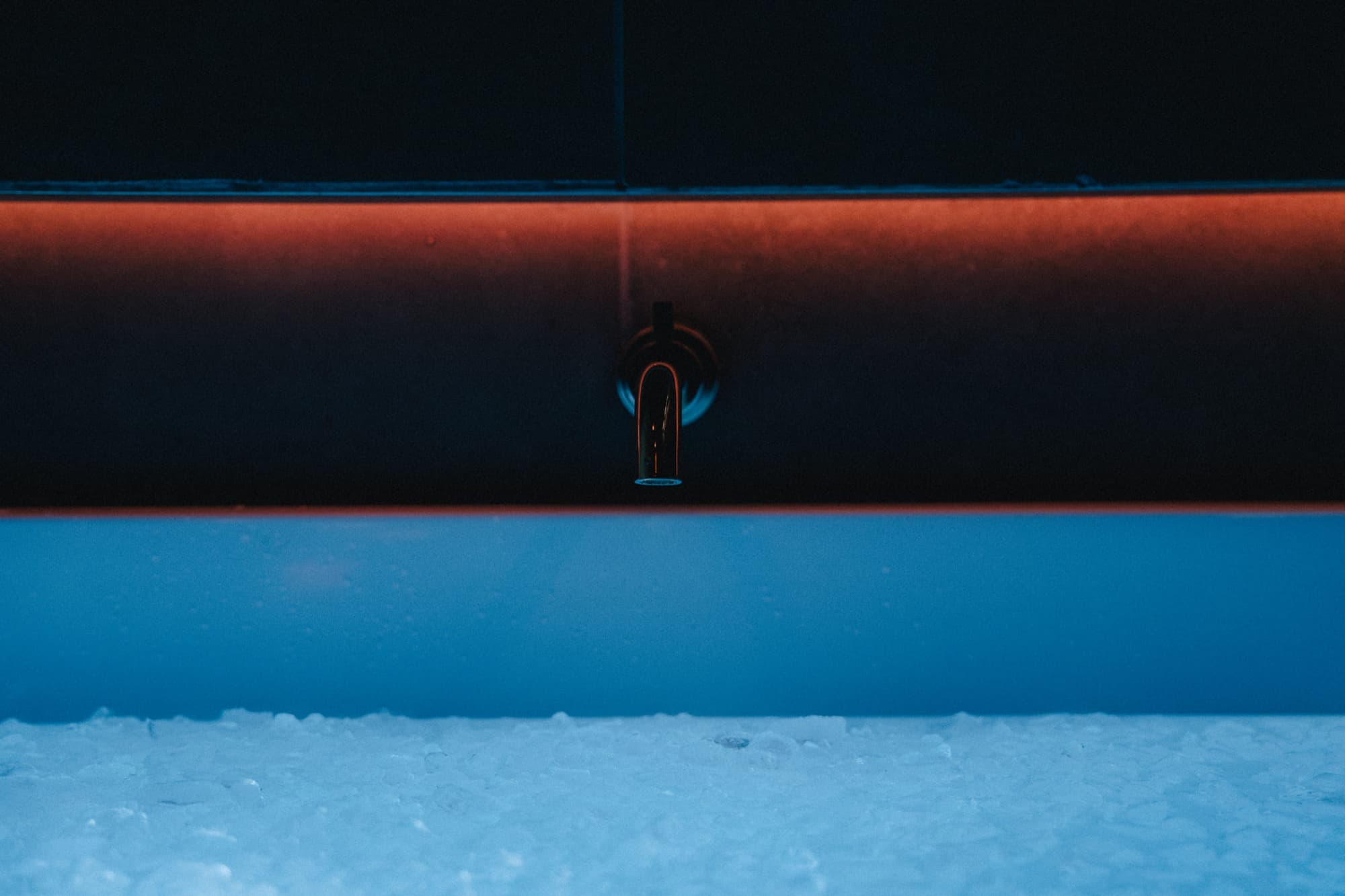 The ice bath's brass spout under a warm cove light, pale blue water and ice below